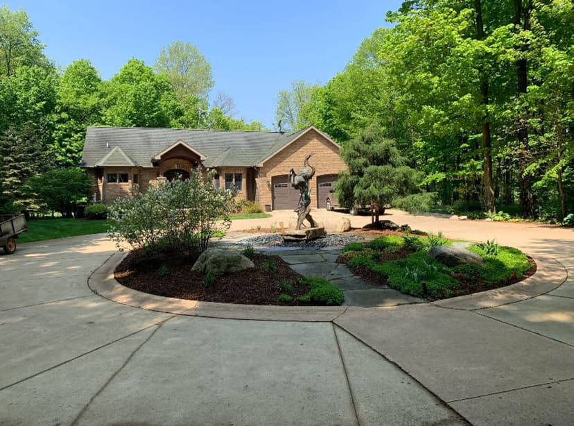 Brick house with landscaped garden and statue in front, surrounded by lush green trees.